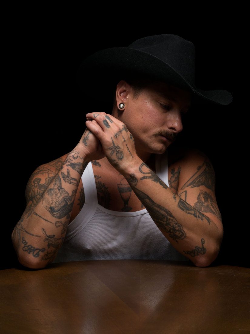 A tattooed man in a black cowboy hat and white tank top leans on a table, hands clasped beneath his chin, looking down in low light; colour.