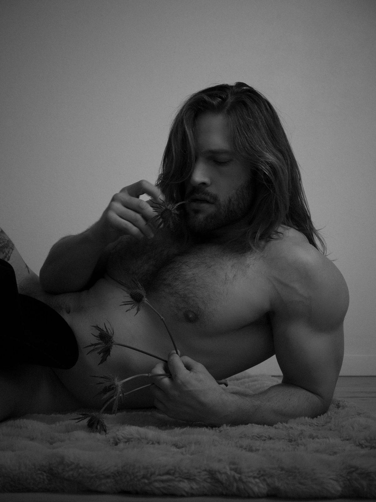A long-haired, bearded man reclines on a fur rug, shirtless, turning a dried flower in his fingers, eyes lowered; black and white.