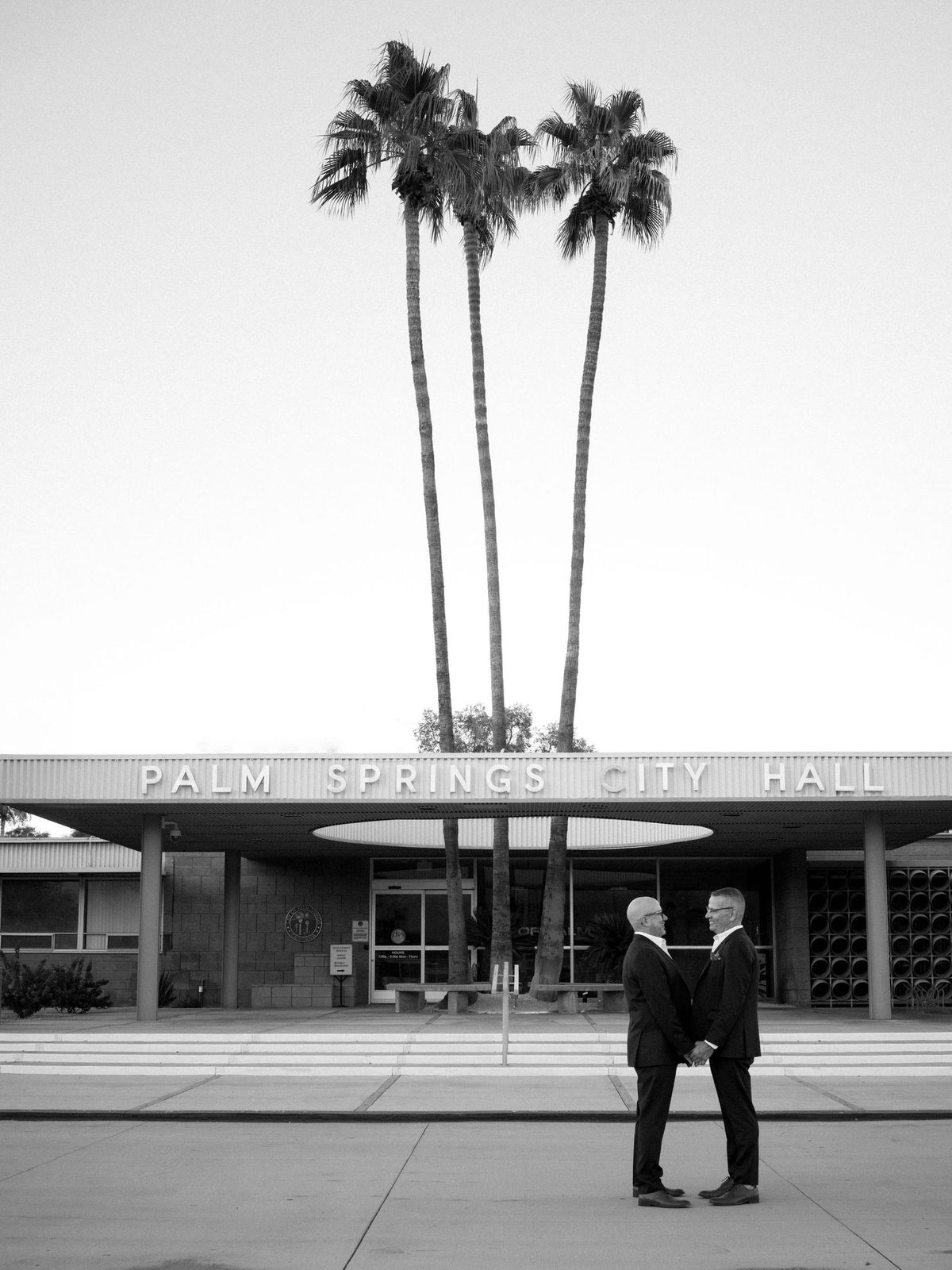 Two men in dark suits stand hand in hand outside a midcentury city hall beneath tall palm trees; black and white.