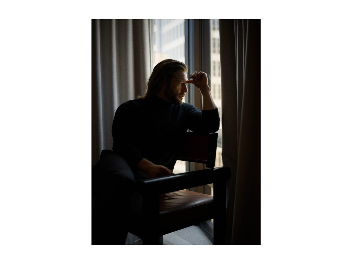 Spread 1 of 15 from An American Pyscho: a long-haired man sits in a dark armchair by a window, head resting on his hand; colour, low light.
