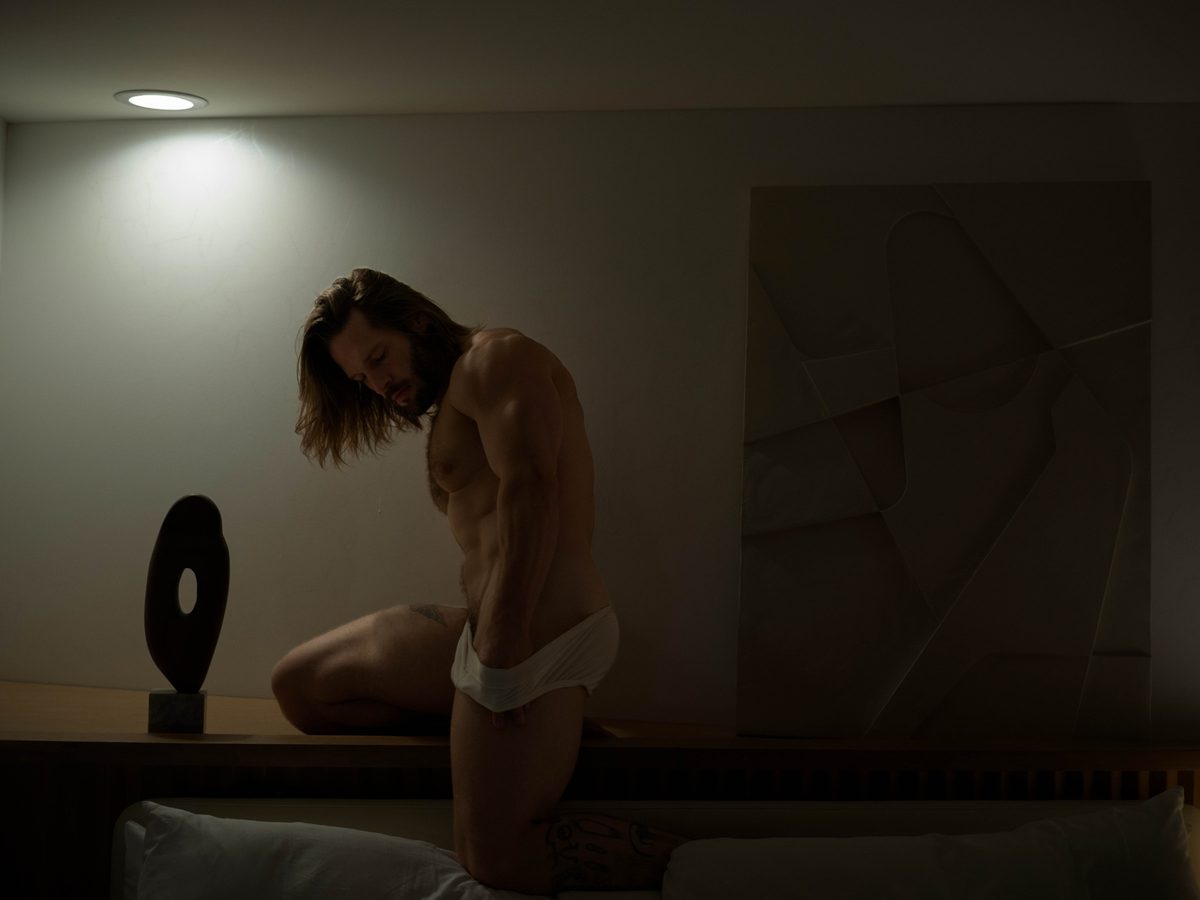 Spread 8 of 15 from An American Pyscho: a long-haired man in white briefs kneels on a low table beside a sculpture, head bowed; colour, low light.