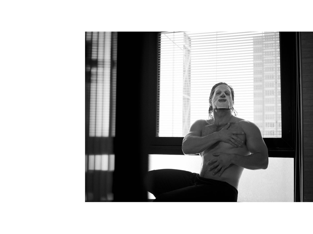 Spread 12 of 15 from An American Pyscho: a long-haired man wearing a white face mask sits bare-chested at a window, arms crossed; black and white.