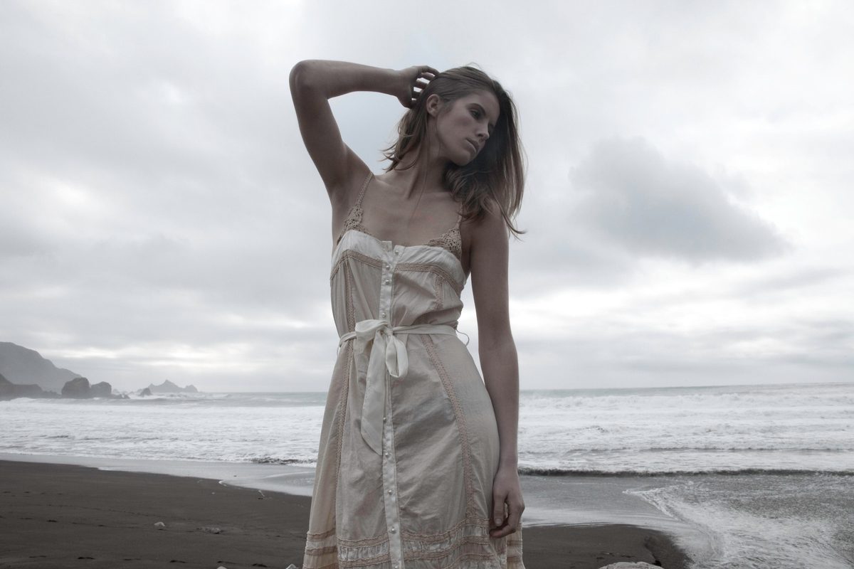 Spread 1 of 3 from Pacifica Romantica: a woman in a pale slip dress stands on a dark sand beach, wind in her hair; colour, overcast.