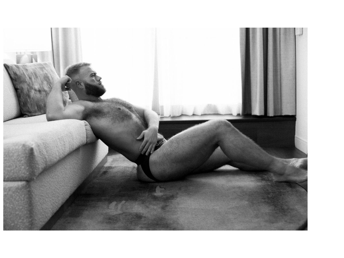 Spread 1 of 4 from A Quiet Confidence: a bearded man reclines on the floor against a sofa, wearing a jockstrap, legs stretched out; black and white.