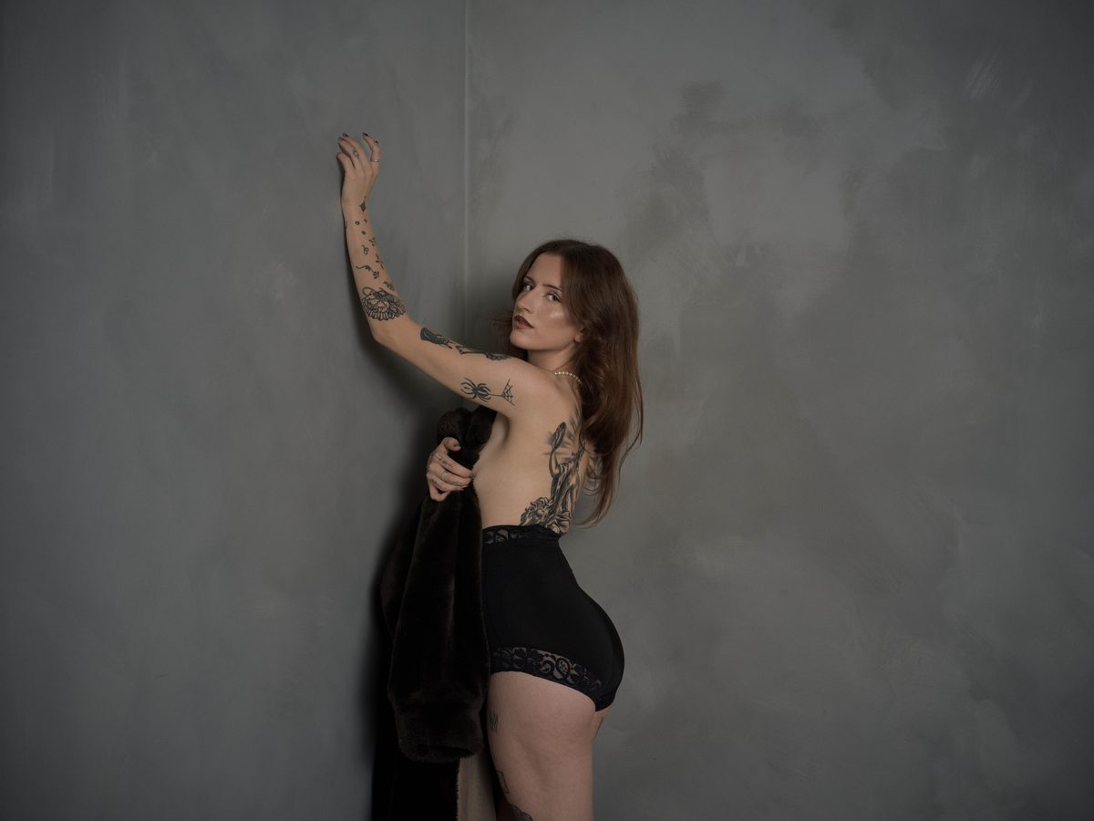 Spread 1 of 7 from The Apprentice: a tattooed woman in black briefs leans against a wall, one arm raised, a dark coat draped from her other hand; black and white.