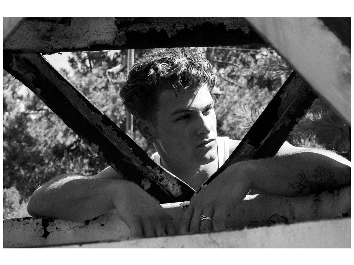 Spread 9 of 12 from On The Road: a young man in a tank top leans through a rusted metal frame outdoors, tattoos on his forearm; black and white.