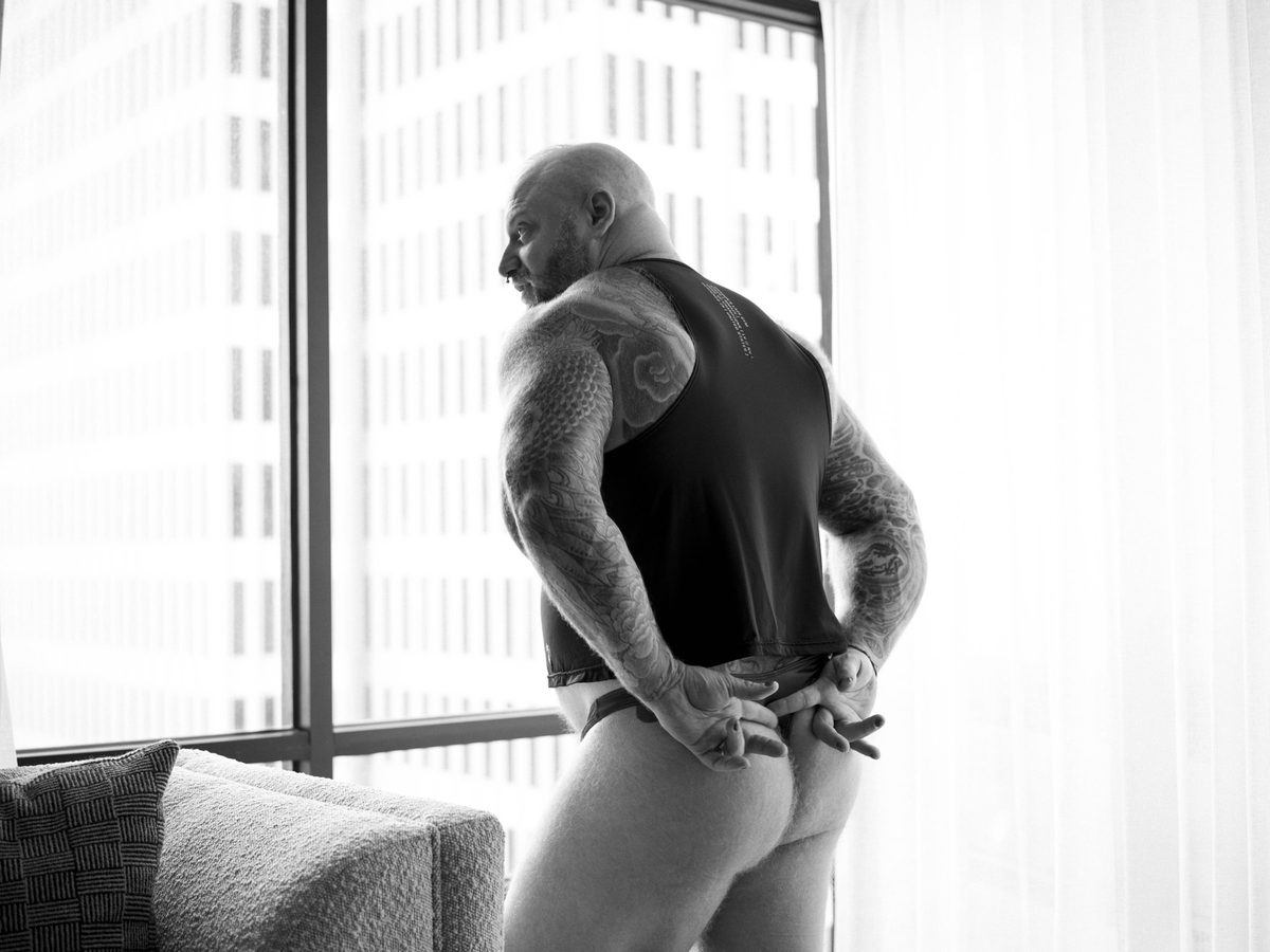 Spread 1 of 8 from Beef, Brawn and Bare: a heavily tattooed, bald man stands at a window in a tank top, pulling down briefs at the hip; black and white.