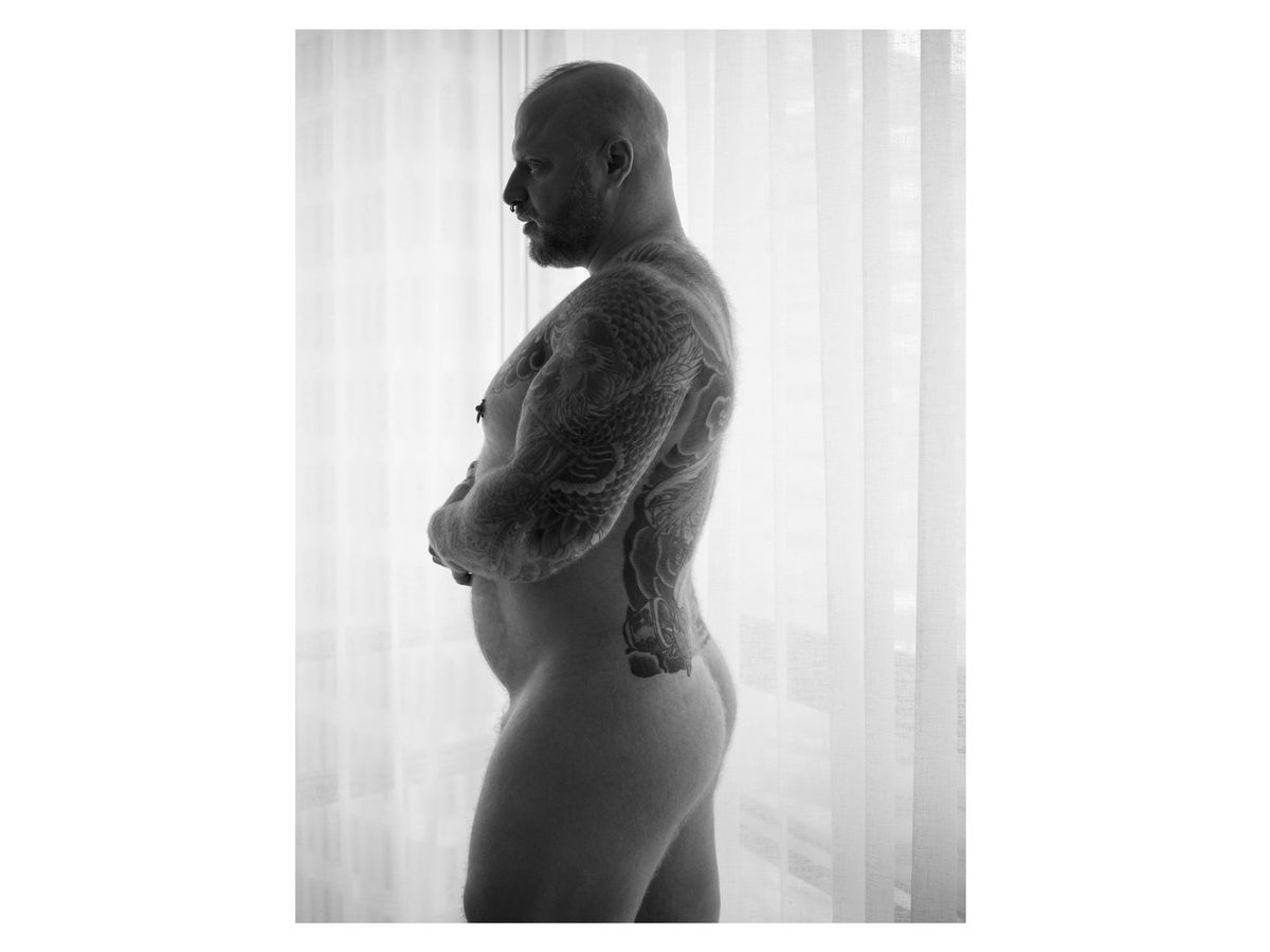 Spread 5 of 8 from Beef, Brawn and Bare: a heavily tattooed, bald nude man stands in profile at a curtained window, arms folded; black and white.