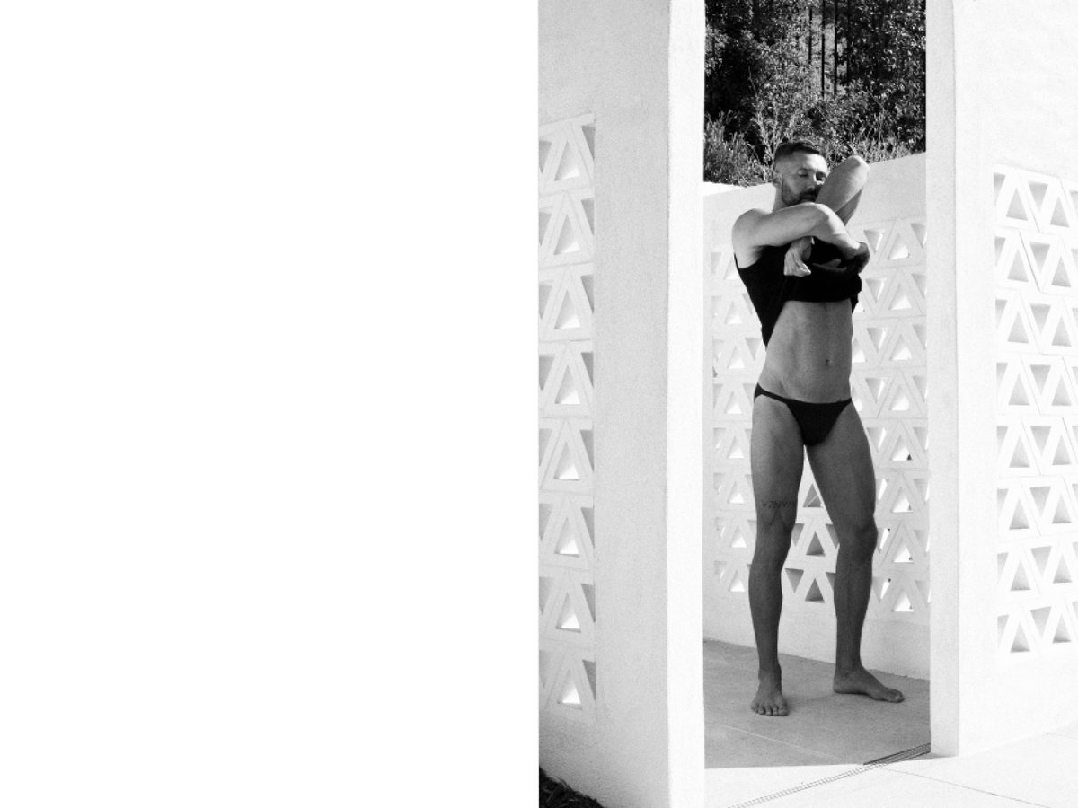 Spread 1 of 2 from Beverly Hills: a man in a black top and briefs stands in a breezeway doorway, pulling the shirt overhead; black and white.