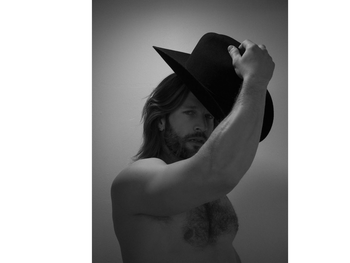 Spread 1 of 10 from Cowboy Daisy: a long-haired, bare-chested man holds a black cowboy hat up beside his face; black and white.