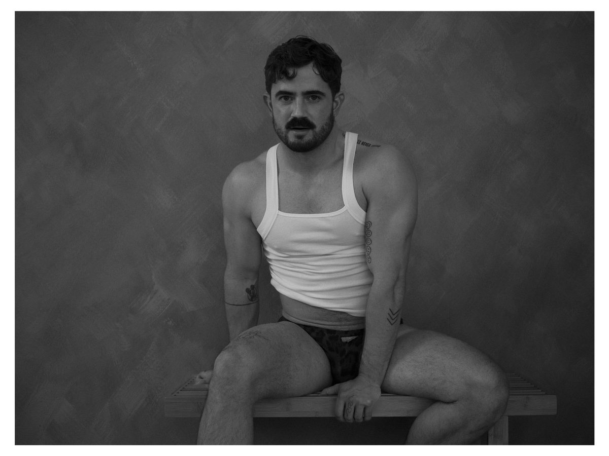 Spread 8 of 11 from Good Boy Supply: a moustached man in a tank top and briefs sits on a wooden bench against a textured wall; black and white.