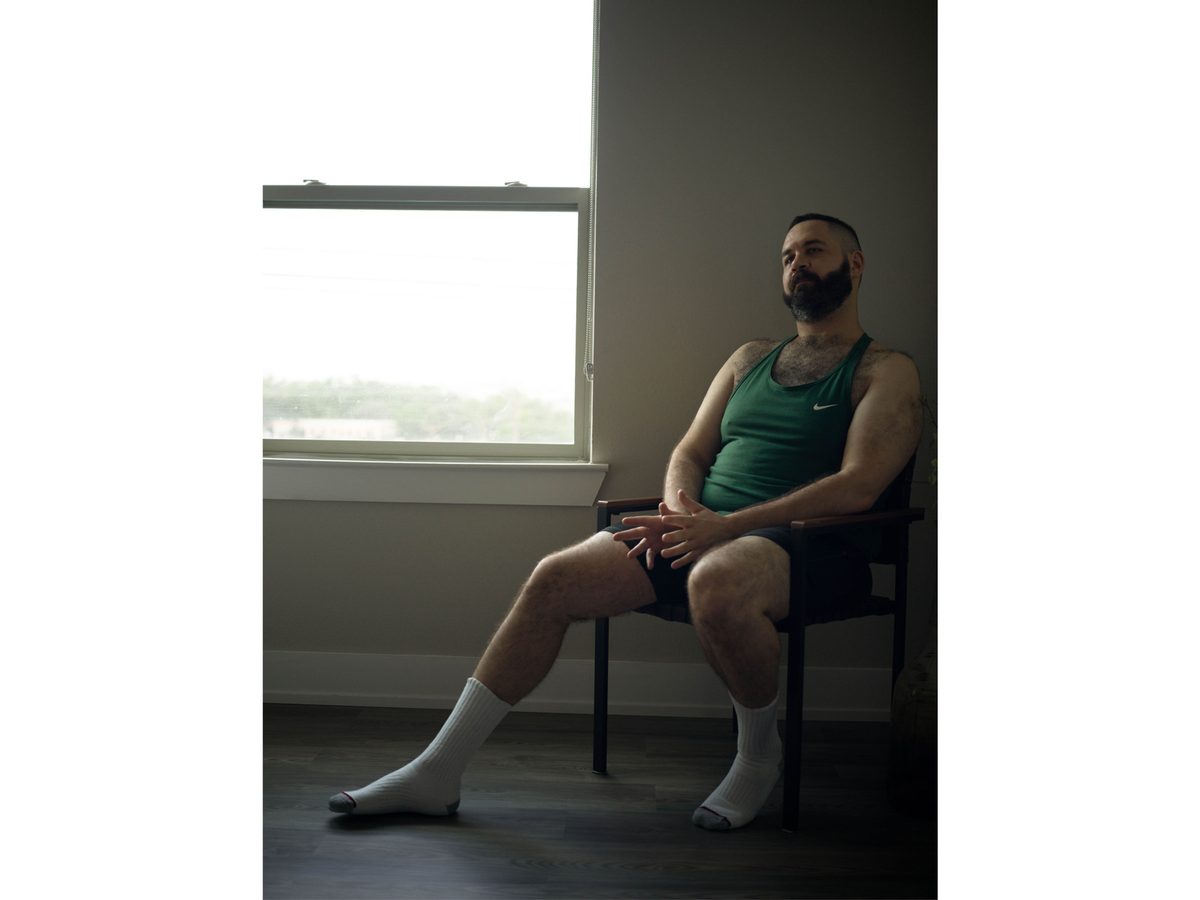 Spread 1 of 2 from Soft Purity: a bearded man in a green tank top sits by a window, legs crossed, looking aside; colour.
