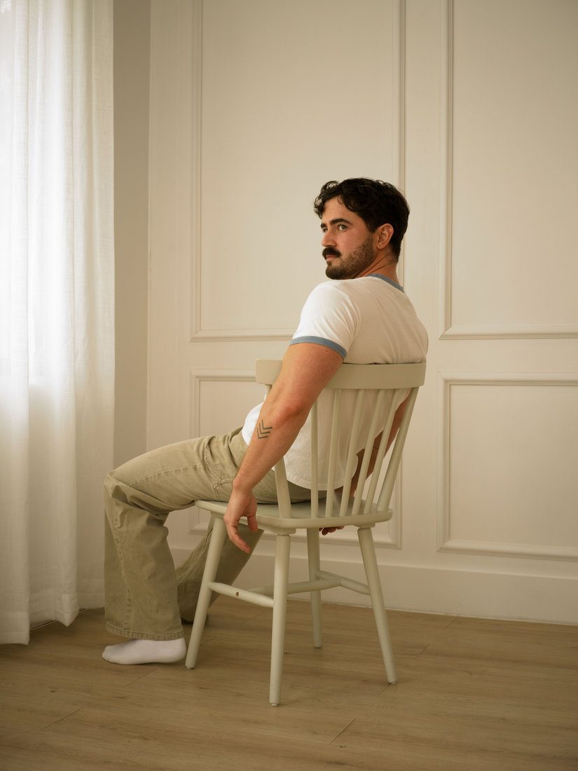 A man in a white t-shirt and tan trousers sits backward on a wooden chair, looking off to the side; colour.