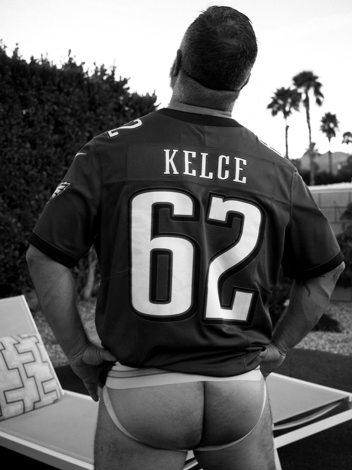 A man in a sports jersey and jockstrap stands outdoors among palm trees, seen from behind; black and white.