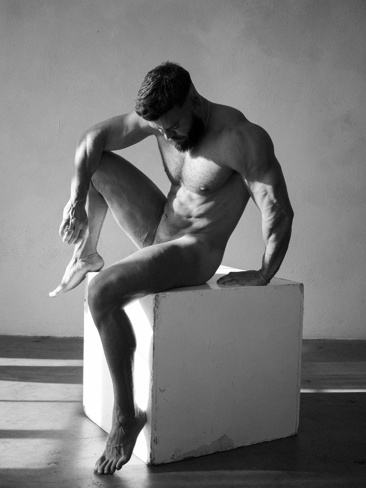 A bearded, muscular nude man sits on a white cube, one leg raised, head bowed in profile; black and white.