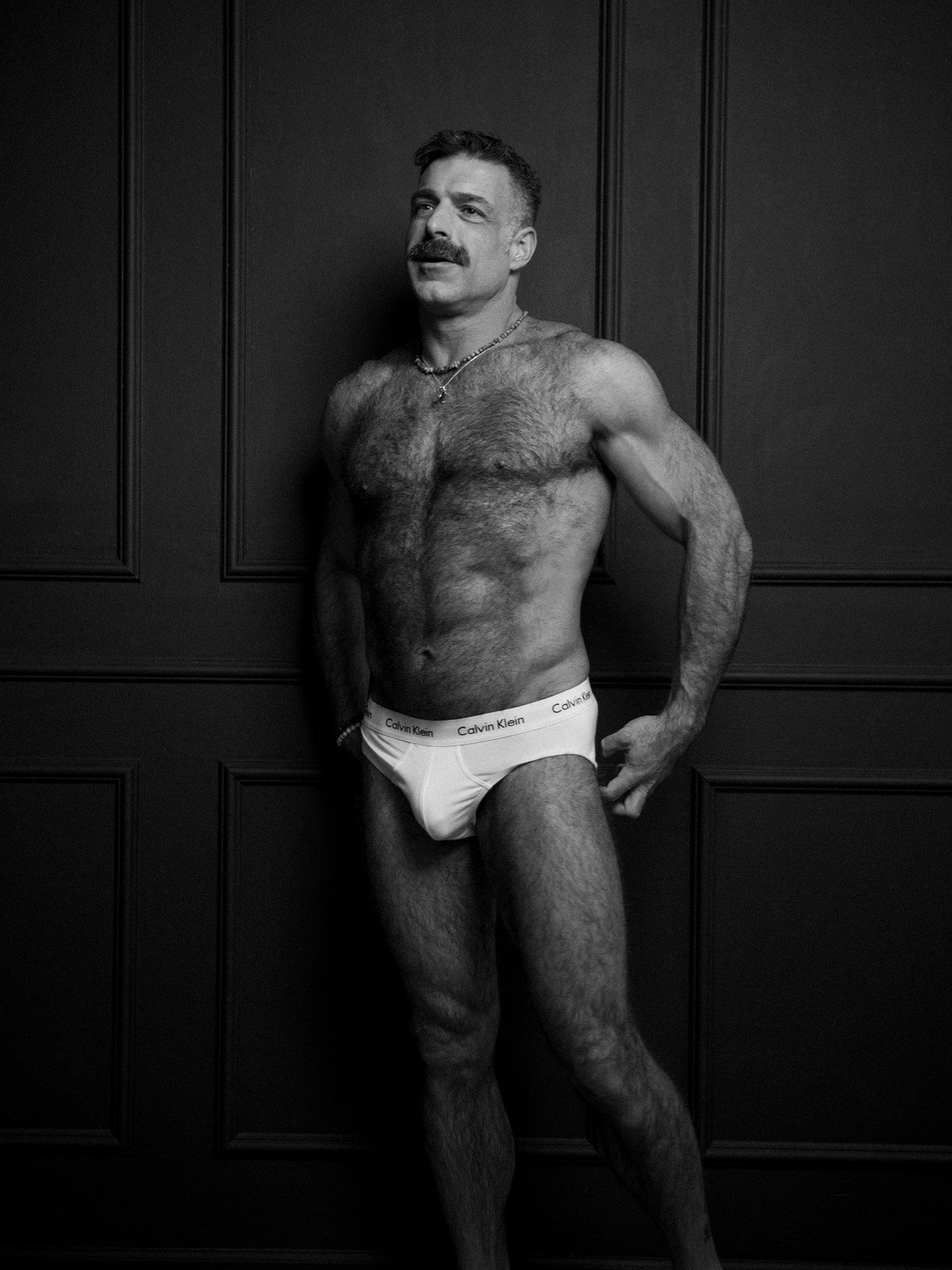 A bearded, hairy-chested man in white briefs leans against a dark panelled wall, one hand on his hip; black and white.