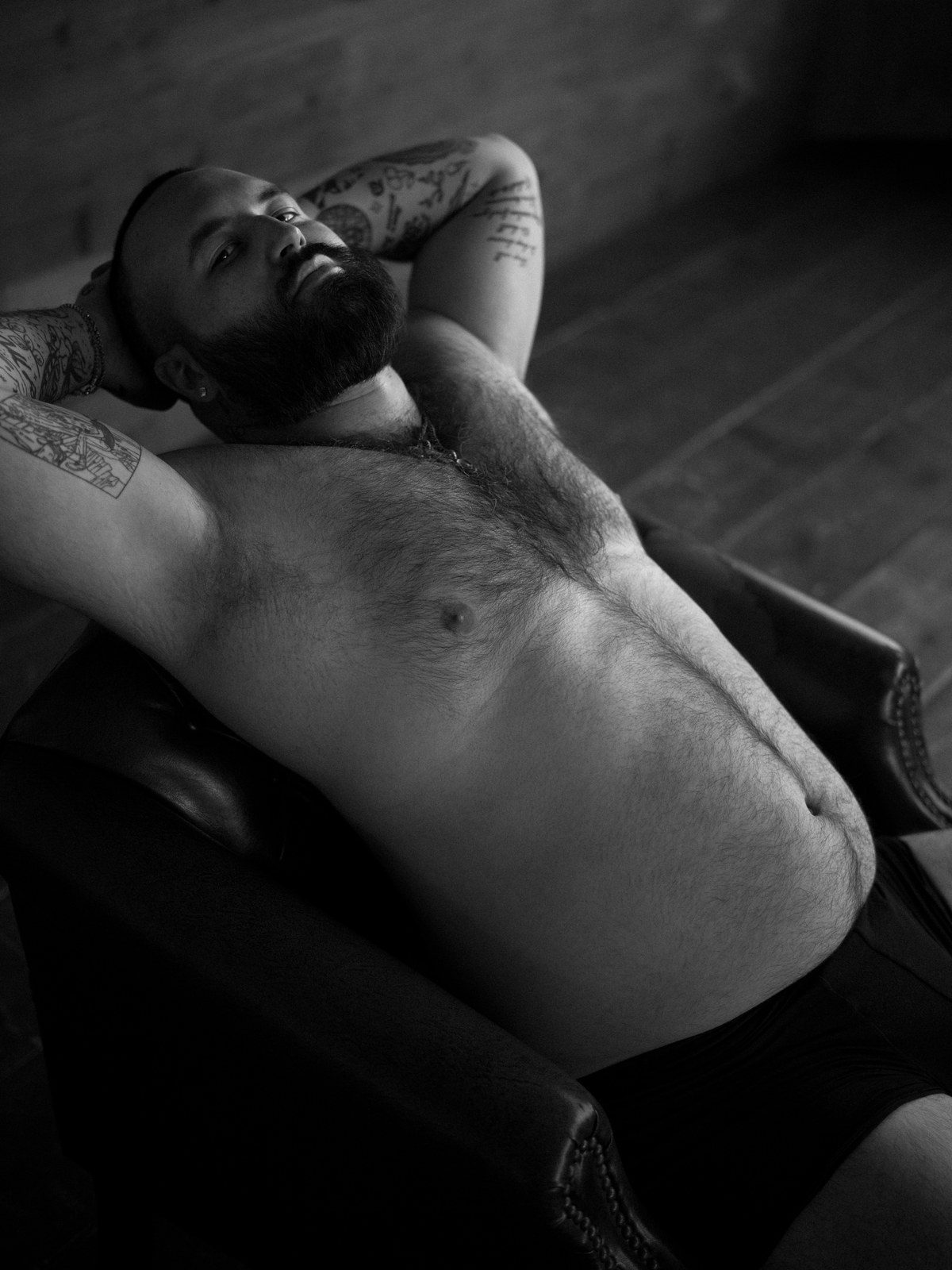 A bearded, tattooed, heavyset man reclines shirtless with one arm behind his head, eyes closed in dim light; black and white.