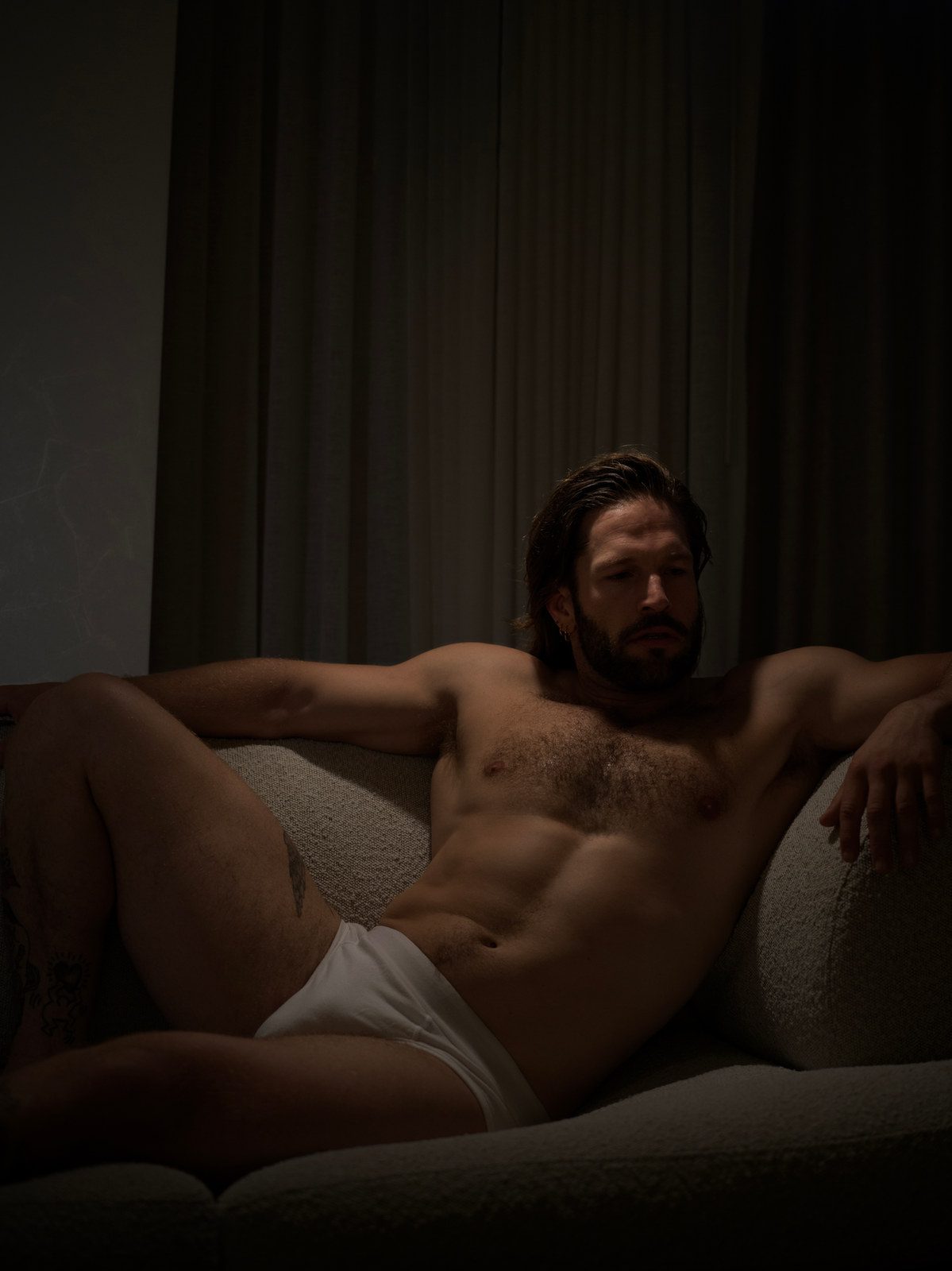 A bearded, shirtless man reclines on a sofa in white briefs, head resting against the cushions; black and white, dim light.