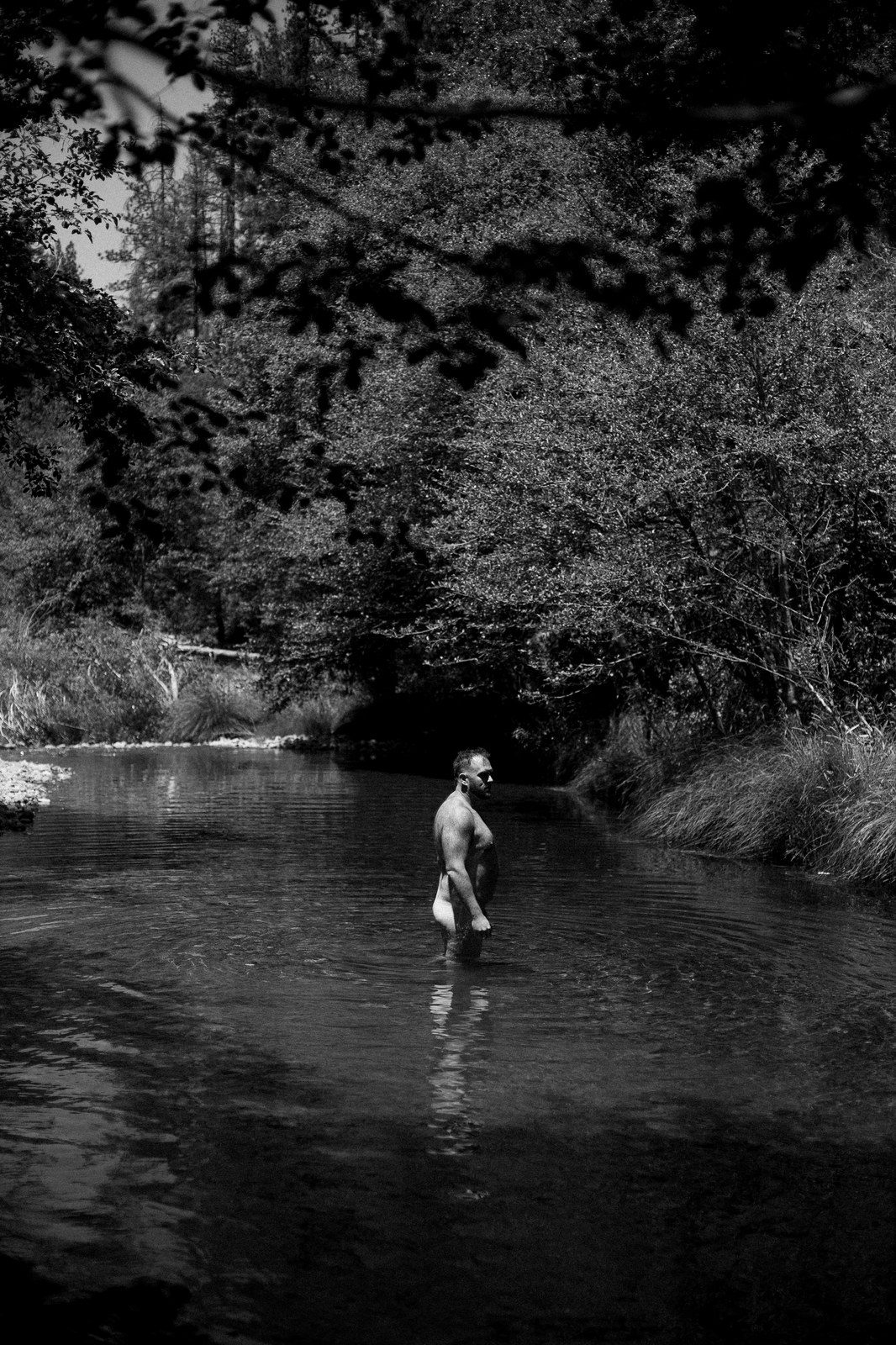 A man wades waist-deep in a wooded stream, shirtless, seen at a distance among overhanging trees; black and white.