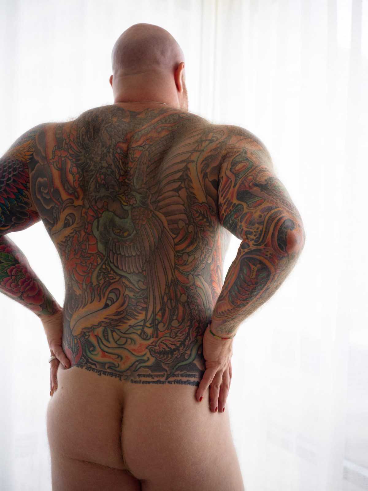 A bald, heavily tattooed man stands at a curtained window, seen from behind, hands on his hips; colour, backlit.