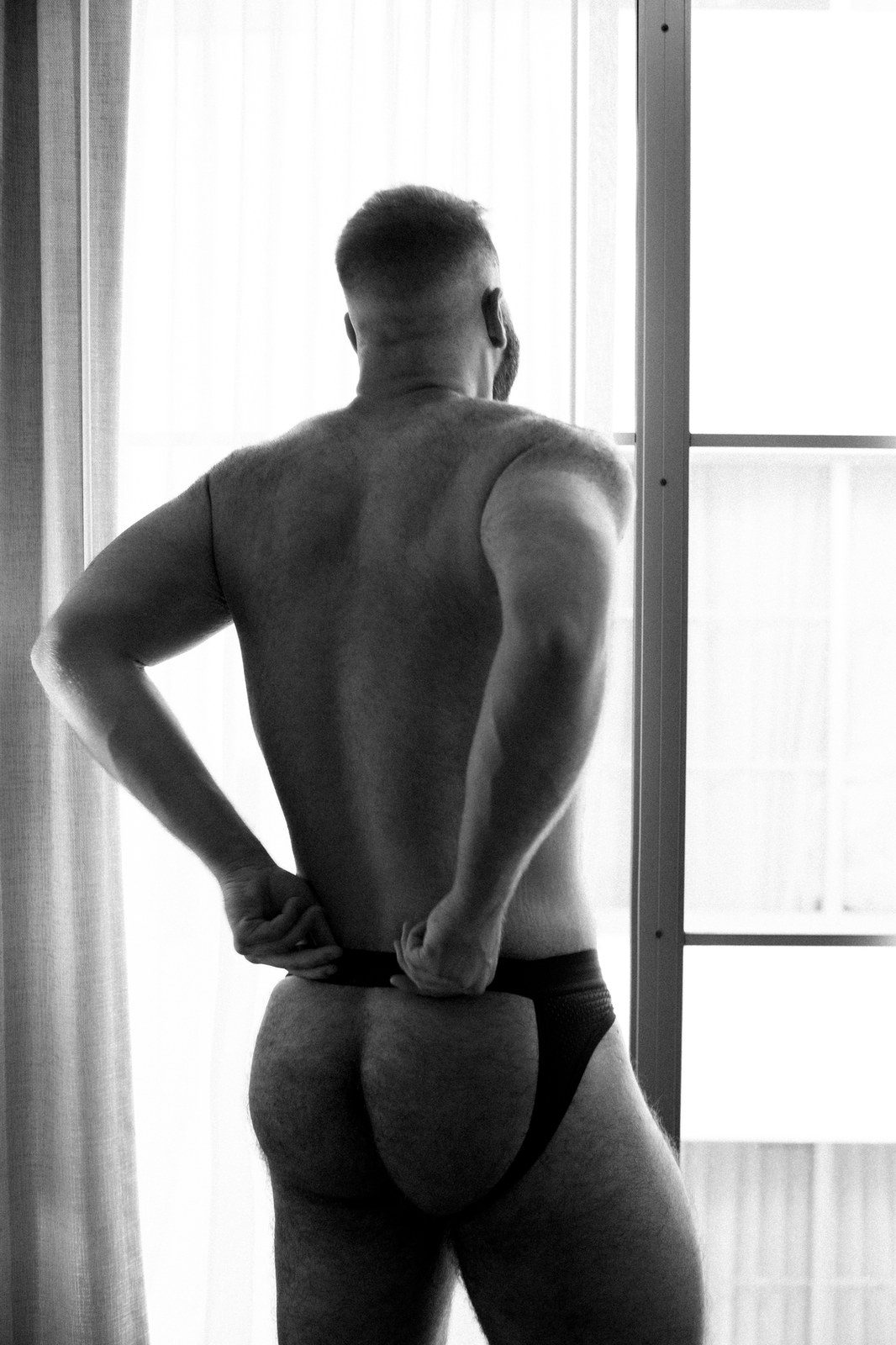 A man in a jockstrap stands at a sunlit window, seen from behind, adjusting the waistband; black and white.