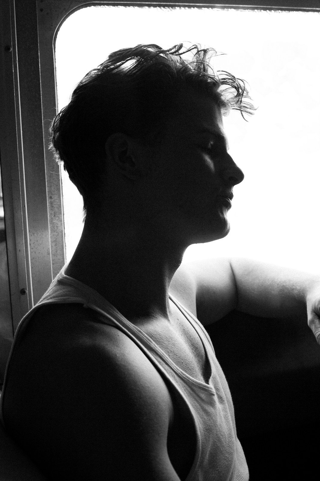 A man in a tank top stands in profile at a bright window, backlit with tousled hair; black and white.