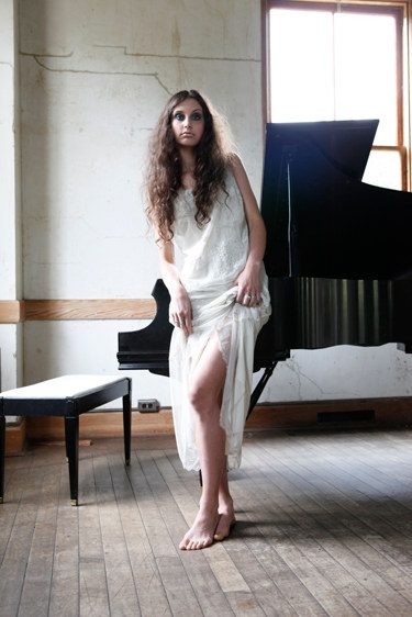 A woman in a pale dress stands beside a grand piano in a bare, sunlit room, looking at the camera; colour.