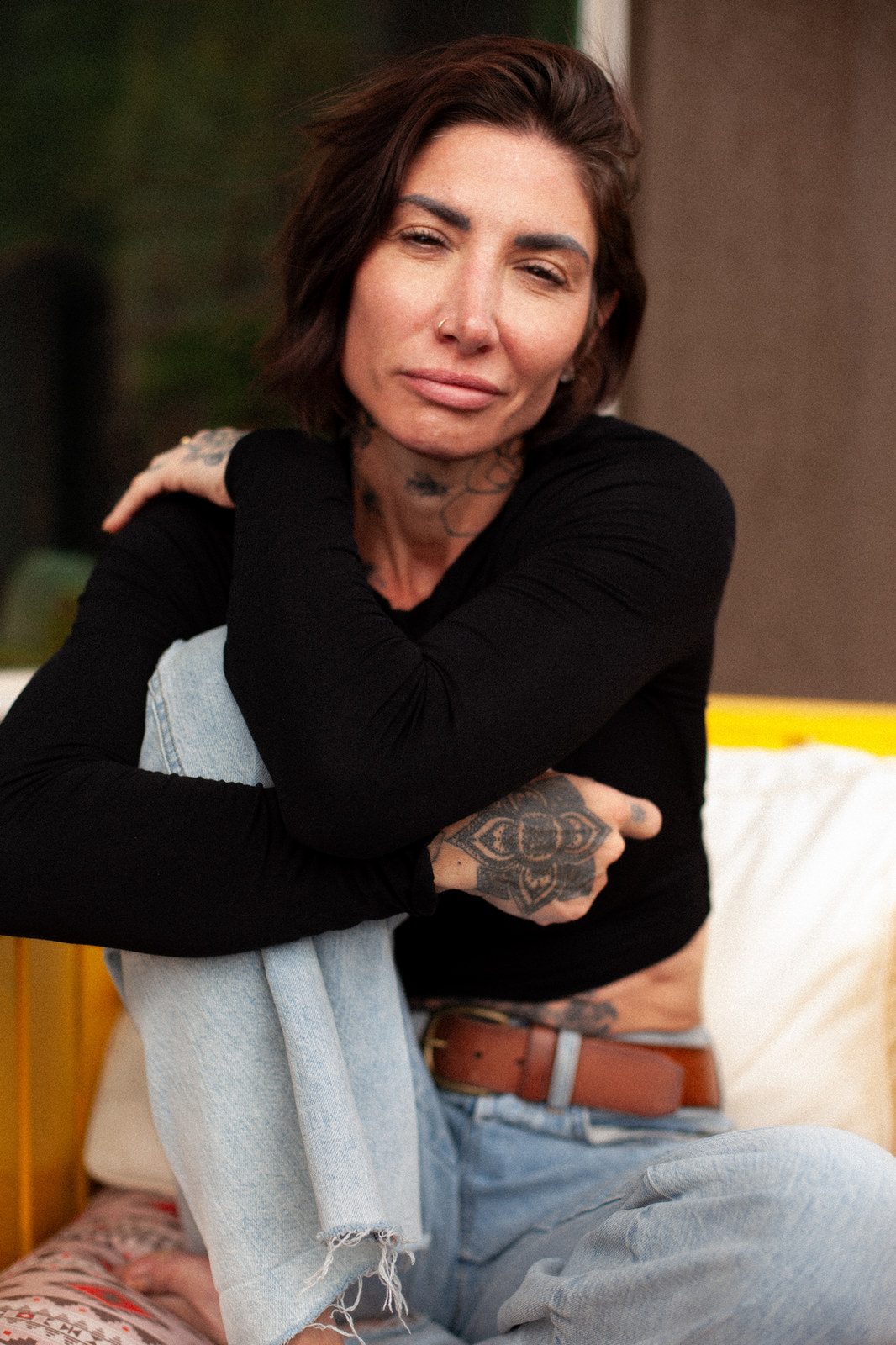 A woman with tattooed hands sits wrapped in a scarf, arms crossed, looking at the camera; colour.