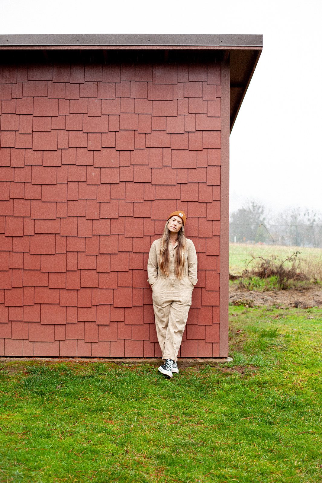 A woman in a tan jumpsuit and cap leans against a shingled wall in a grassy field; colour.
