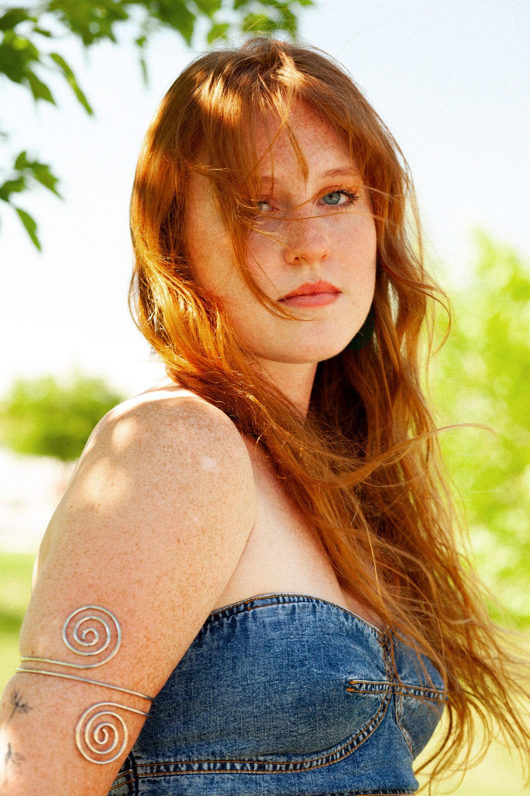 A close portrait of a red-haired woman in a denim top, hair blown across her face, outdoors; colour.